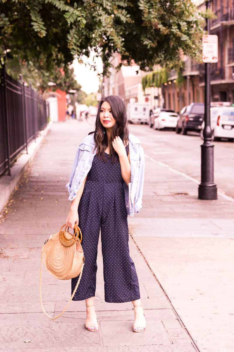 Just A Tina Bit fashion blogger wearing a navy polka dot jumpsuit, denim jacket, and round straw bag in New Orleans