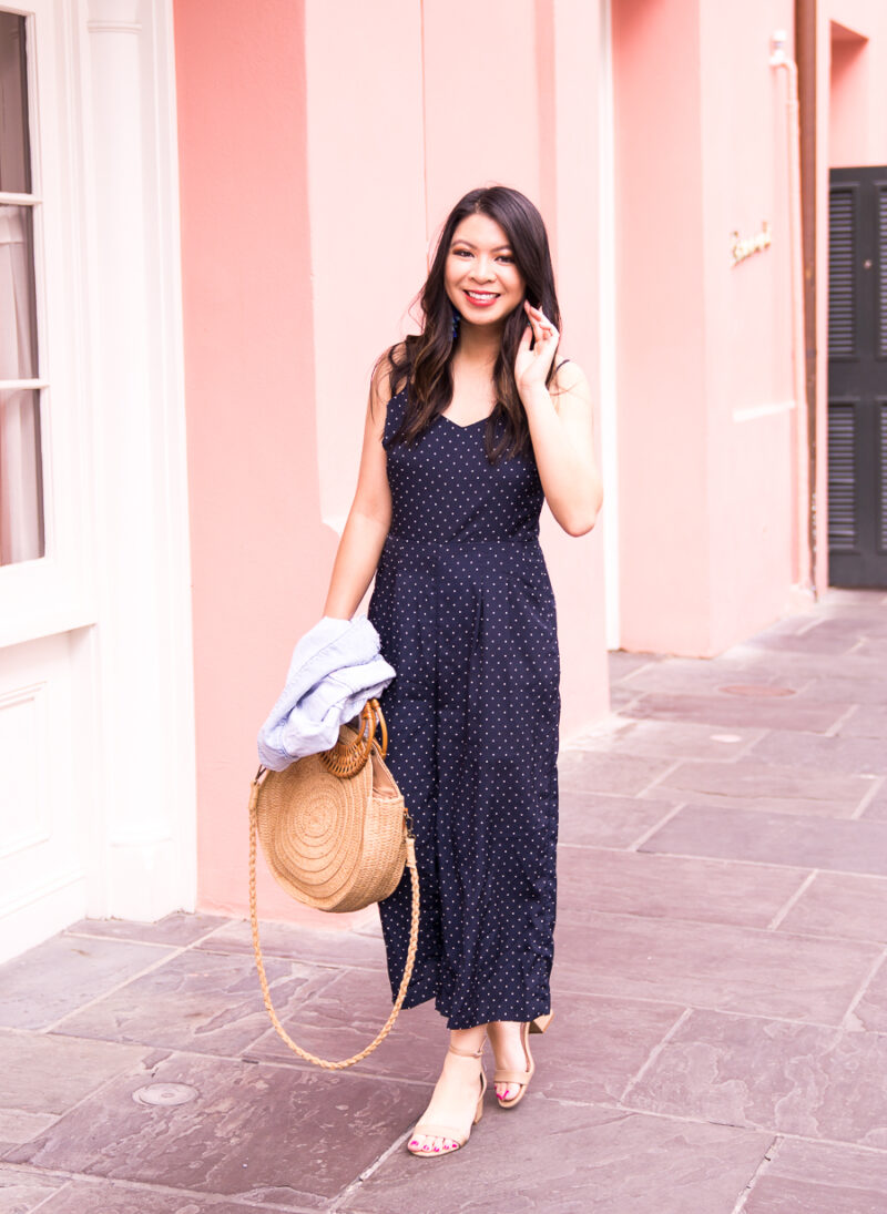 Just A Tina Bit fashion blogger wearing a navy polka dot jumpsuit, denim jacket, and round straw bag in New Orleans, charming pink wall in the French Quarter