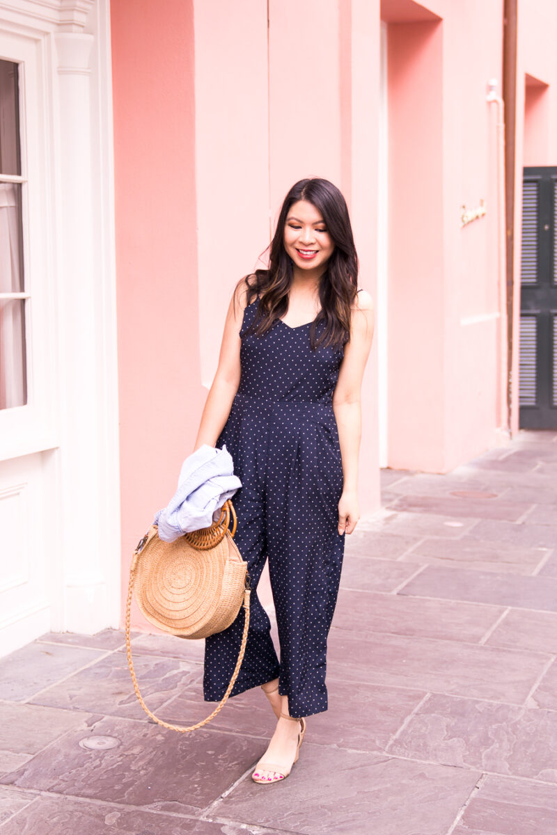 Just A Tina Bit fashion blogger wearing a navy polka dot jumpsuit, denim jacket, and round straw bag in New Orleans, charming pink wall in the French Quarter