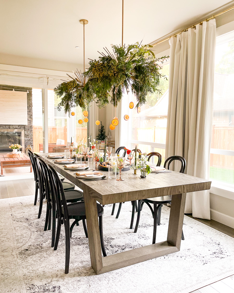 modern holiday tablescape, greenery and dried oranges on dining chandelier, christmas table setting with black chairs and modern wooden table
