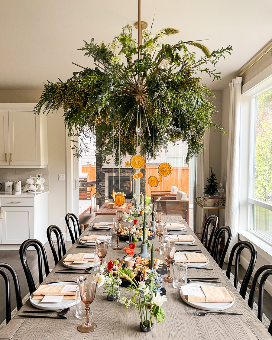 modern holiday tablescape, christmas table setting with black silverware and chairs, greenery and dried oranges on dining chandelier