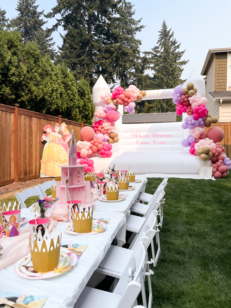 Disney princess birthday party decorations with white bounce house, pink and purple balloon garland, princess cardboard cutout, kids table setting with crown bowls, princess plates, and castle cupcake holder