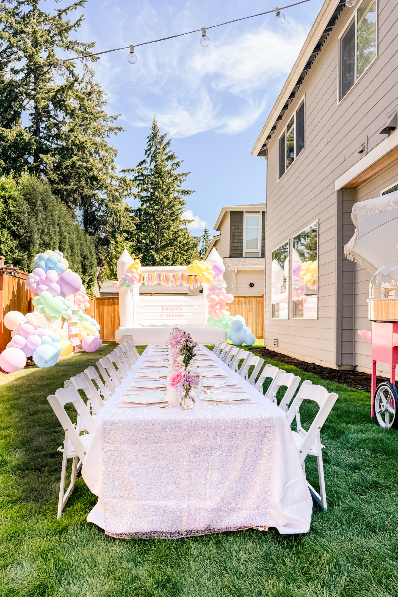 Unicorn birthday party with iridescent table cloth on the kids table, a white bounce house, and pastel and rainbow balloon garland