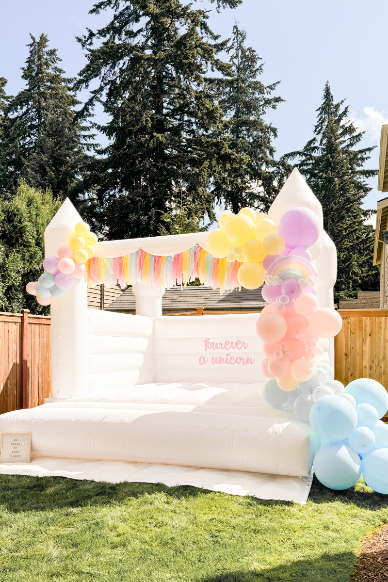 White bounce house with pastel tassels and rainbow balloon garland, DIY decal using smart removable vinyl that fourever a unicorn