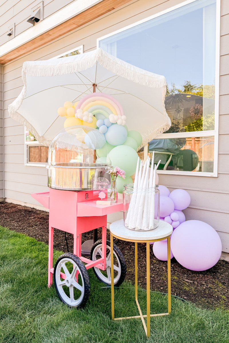 Pink cotton candy machine with cones, DIY acrylic arch sign, and white umbrella for unicorn birthday party, 